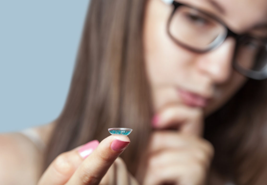 A woman in glasses holds a blue contact lens on her finger